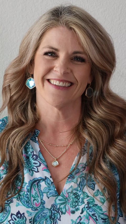 Woman with long wavy blonde hair smiling at camera, wearing floral patterned blouse and silver necklace