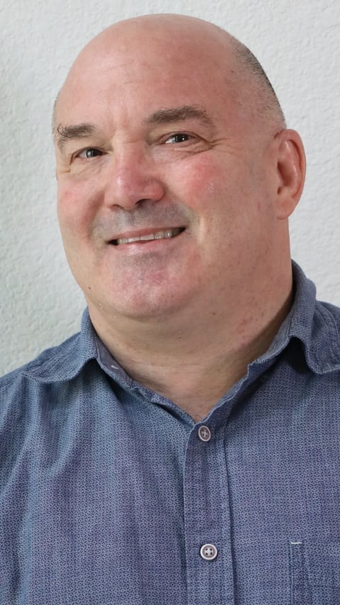Headshot of a middle-aged man with a bald head wearing a blue button-up shirt, smiling at the camera against a white background
