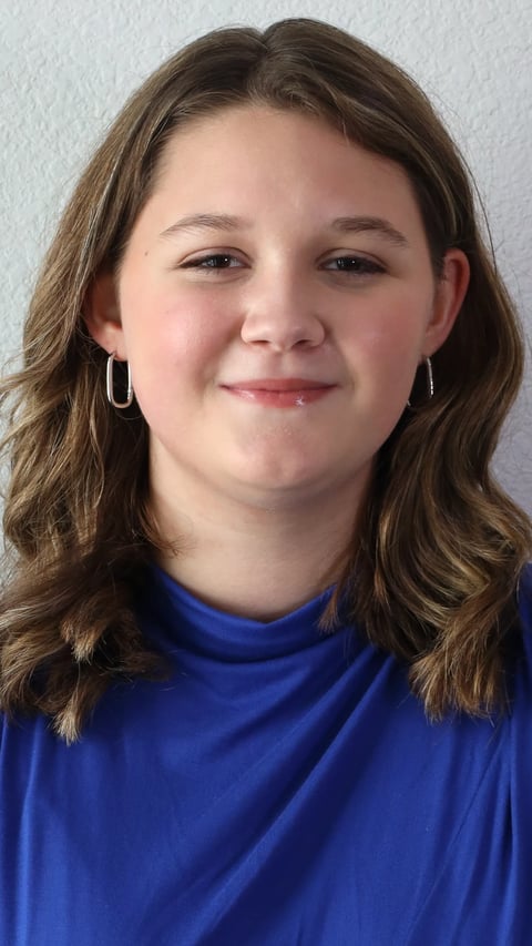 Portrait of a woman with shoulder-length brown wavy hair, wearing silver hoop earrings and a blue turtleneck against a white background.