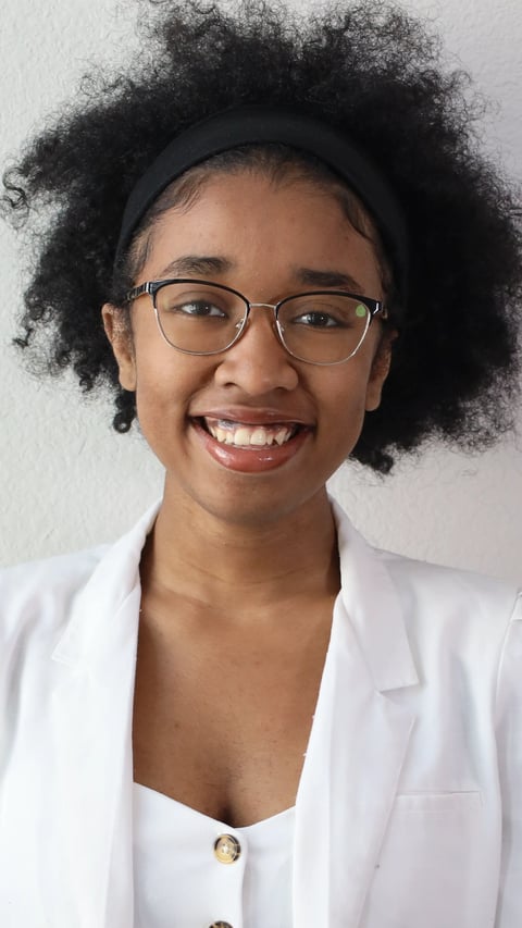 Smiling woman with afro hair, glasses, and black headband wearing a white blazer against a light background