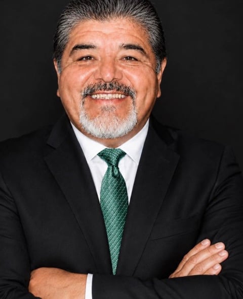 Professional headshot of middle-aged man with gray hair and mustache wearing black suit and dark green tie against black background