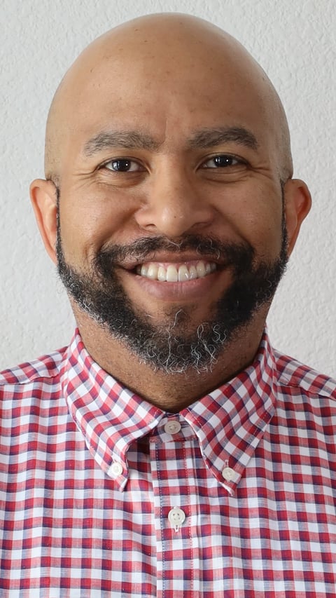 Smiling bald man with dark beard wearing a red and white checkered button-up shirt