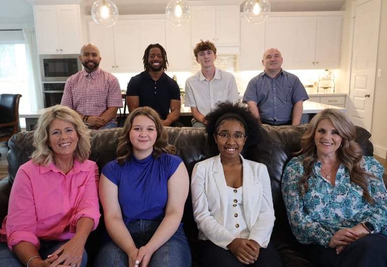 Eight smiling people posing together in a modern kitchen, four standing in back and four seated on a couch in front.